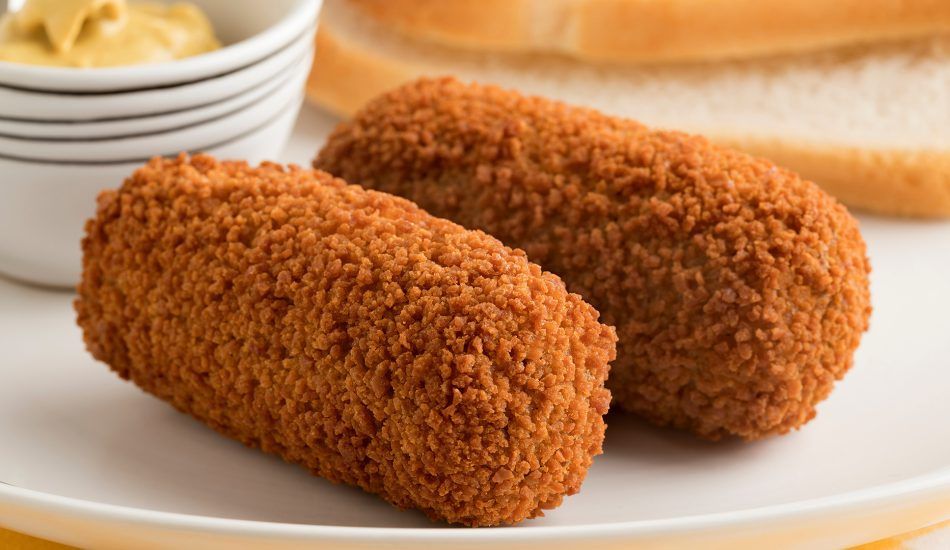 Twee goudbruine, gepaneerde, cilindervormige kroketten op een wit bord met mosterd en brood op de achtergrond.
