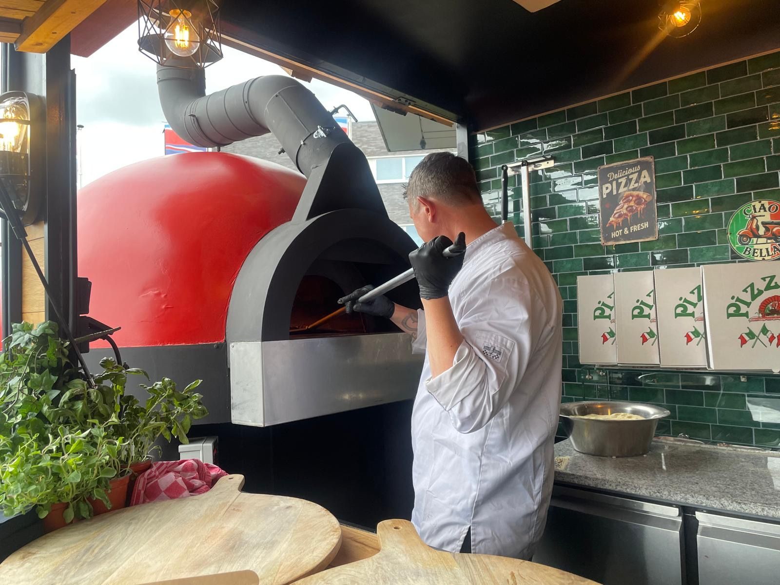 Een chef-kok in een wit overhemd gebruikt een instrument om in een rode pizzaoven te kijken.