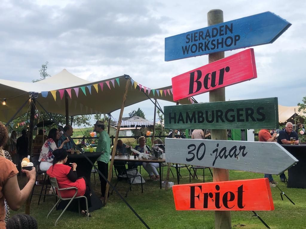Wegwijzer bij een buitenevenement met routebeschrijving: workshop, bar, hamburgers, 30 jaar en friet. Op de achtergrond zijn mensen te zien.