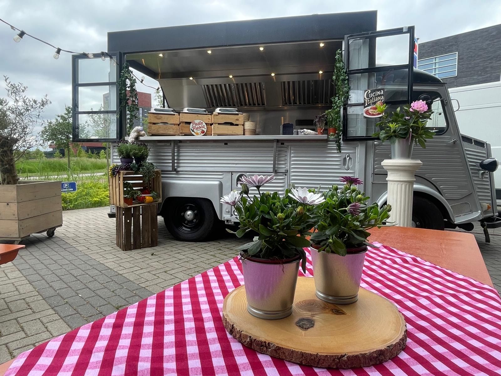 Een foodtruck met open ramen, een roodgeruit tafelkleed en bloempotten op een houten plank.