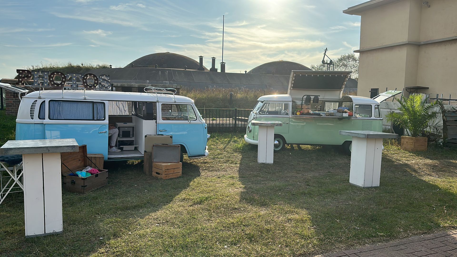 Twee kleurrijke vintage VW-busjes met open deuren en serveertafels op een gazon.
