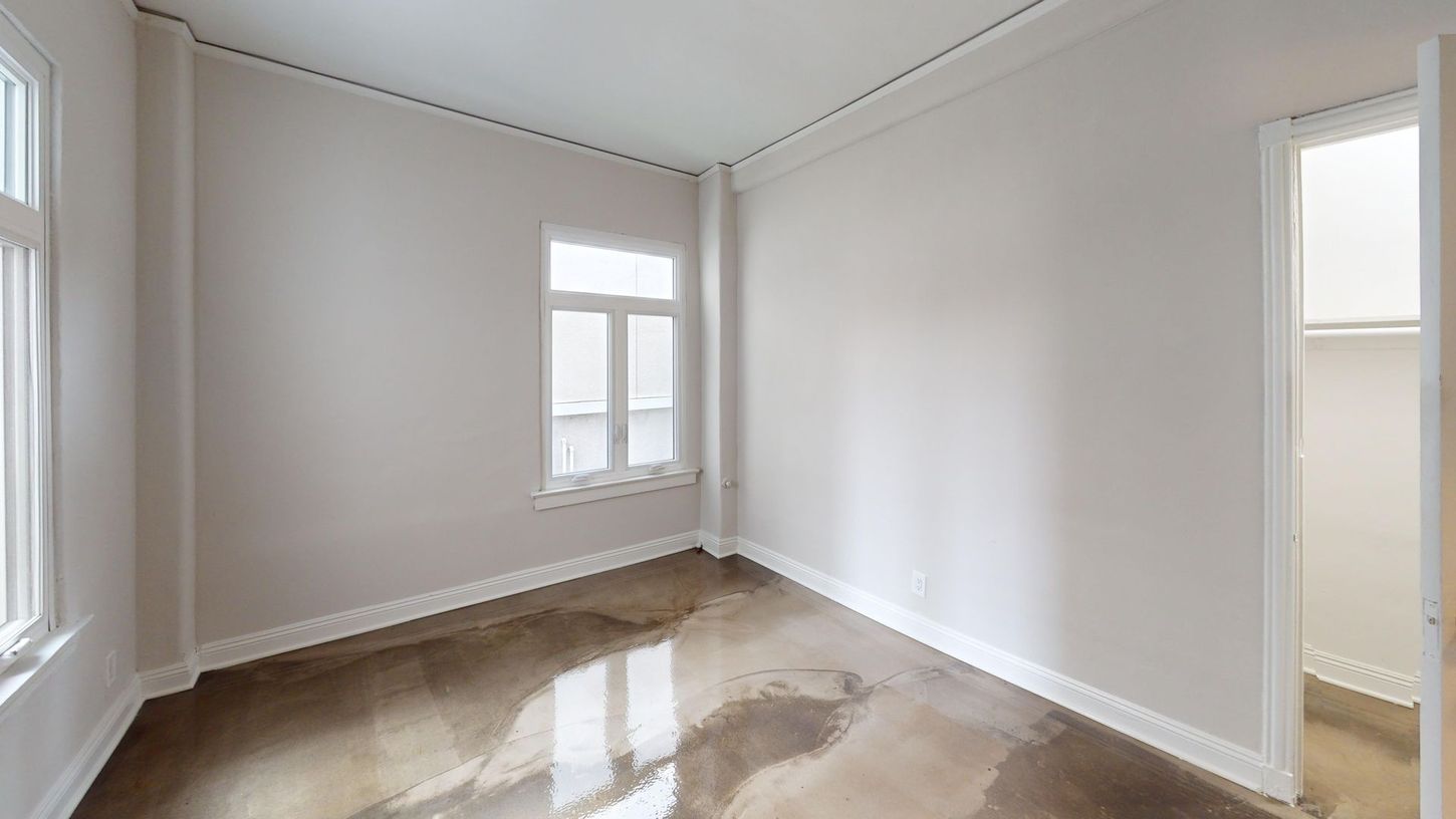 Empty room with white walls, windows, and a concrete floor. A doorway leads to a closet.