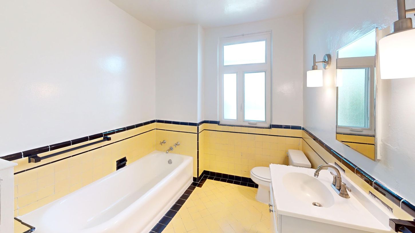 Bathroom with white tub, sink, and toilet; yellow and black tile; window.
