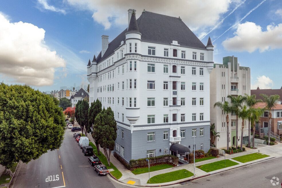 White multi-story apartment building with dark roof, next to a street with cars and trees.