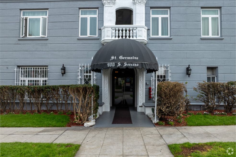 Gray apartment building entrance with black awning, shrubs, and sidewalk.