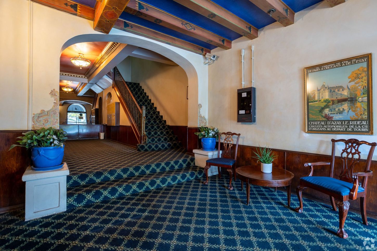 Hallway with ornate details, patterned carpet, stairs, and vintage artwork. Two chairs sit near a small table.