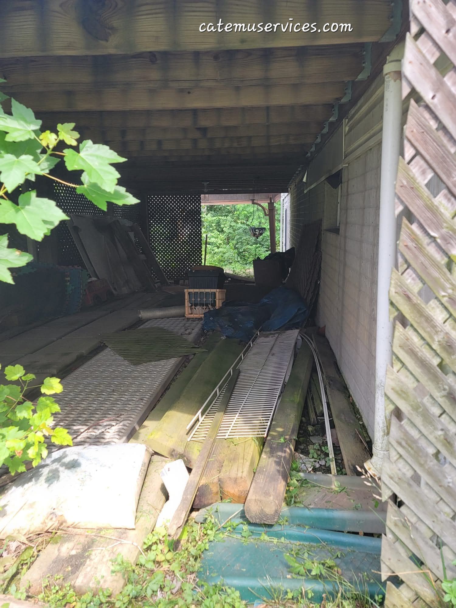 Underneath a wooden deck, a cluttered storage area with various materials, leading to an open doorway with foliage.