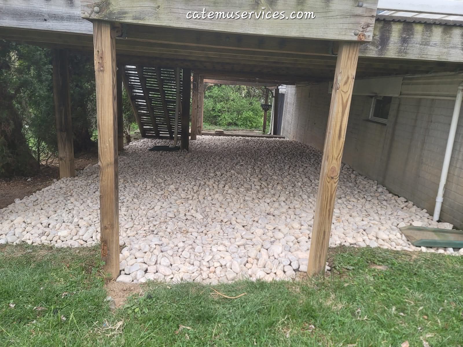 Gravel-covered area under a wooden deck, supported by wooden posts, with stairs and a building in the background.