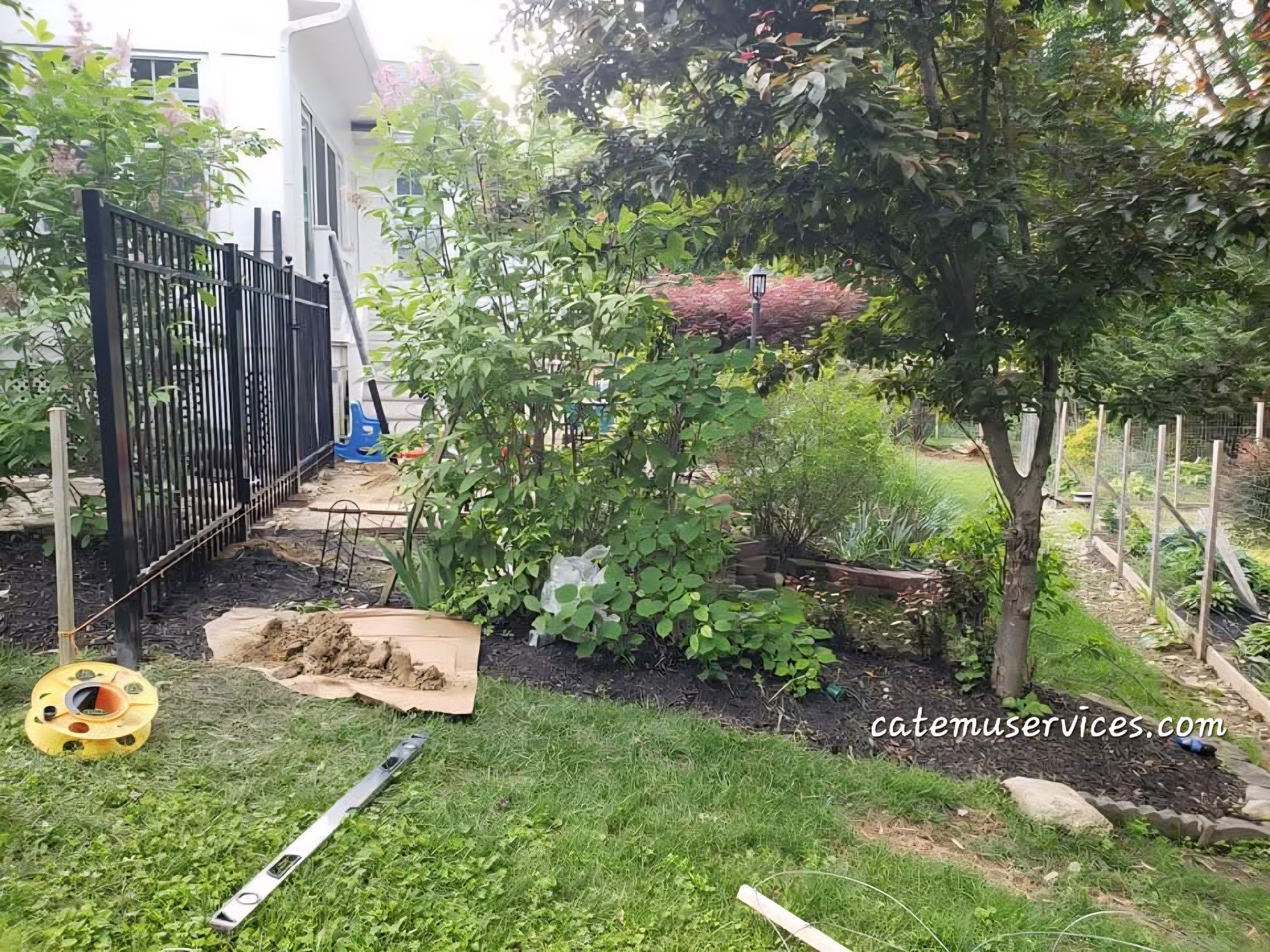 A garden scene with a black fence, tools, and a white house in the background. Lush greenery surrounds the fence.