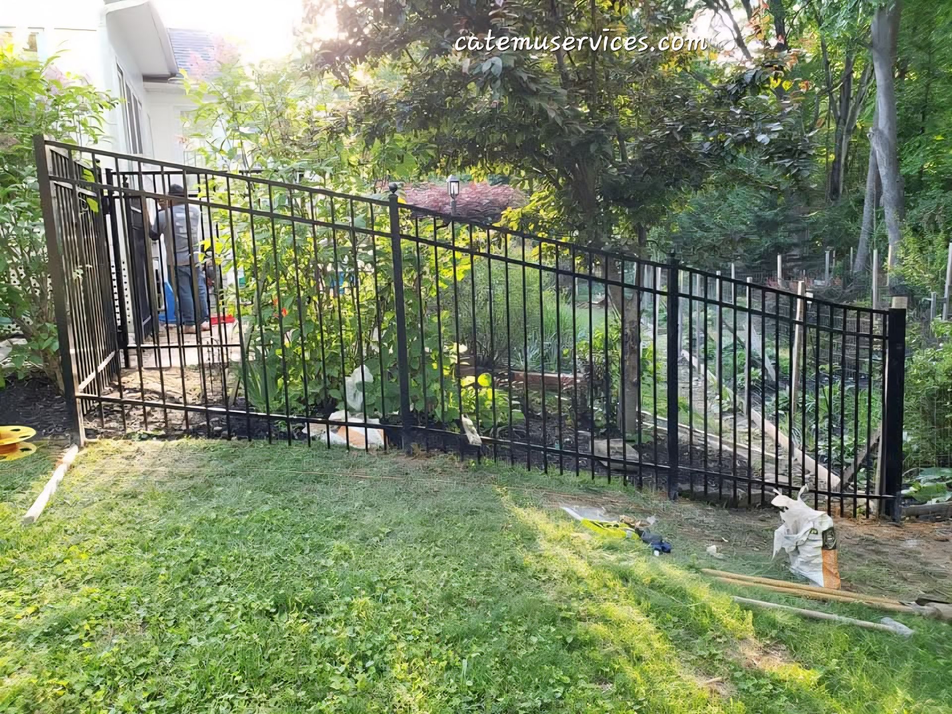 Black metal fence on a grassy hill, surrounding a garden. A person is near the fence.
