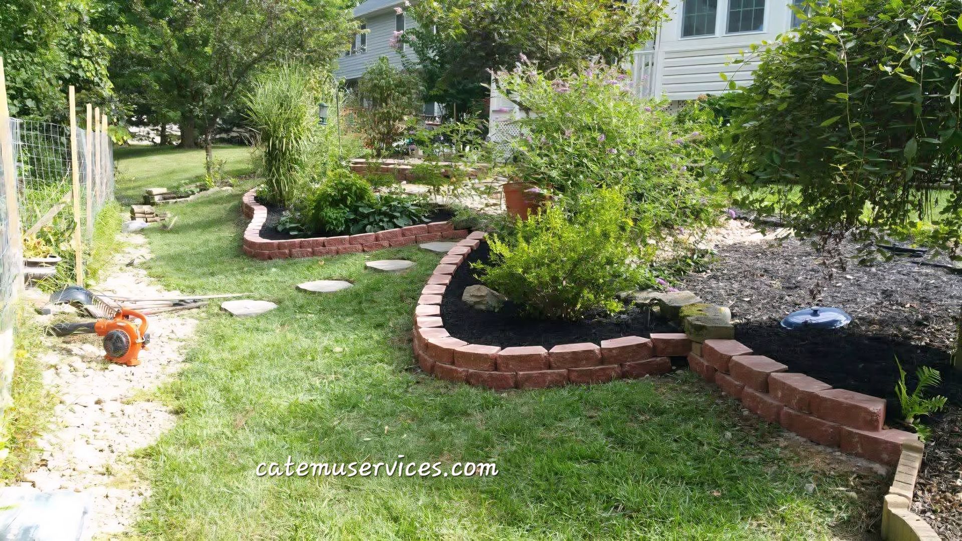 A garden with brick borders, mulch, plants, and stepping stones on a grassy lawn.