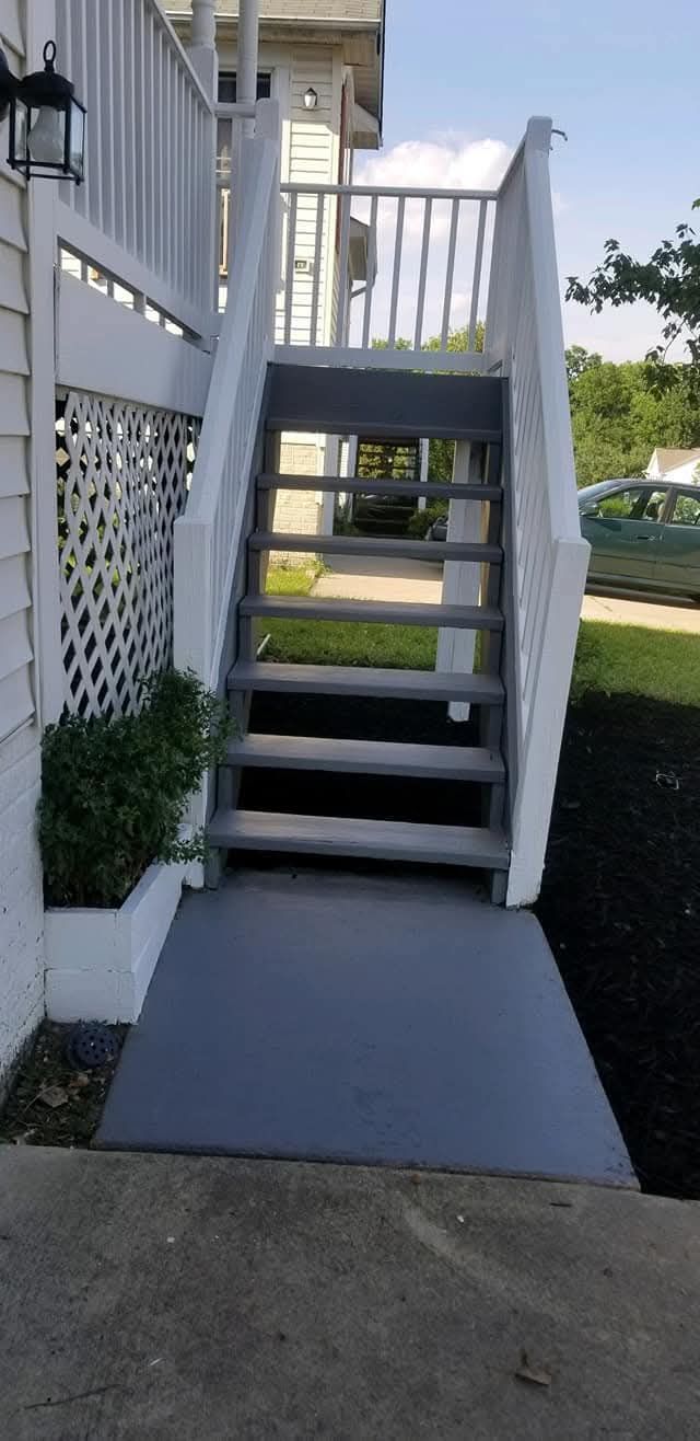 A gray ramp leads to stairs with white railings. A house is on the left, with green shrubbery and a car in the distance.
