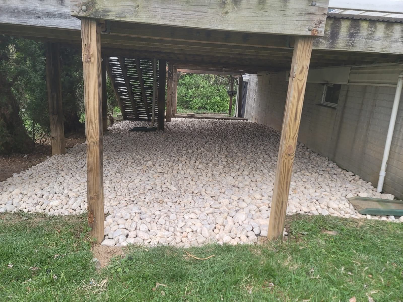 Under a deck, a bed of white stones covers the ground. Wooden posts support the deck. Green grass borders the stones.
