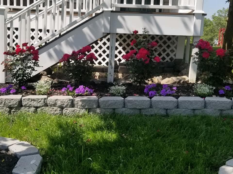 Landscaped garden bed with red roses, purple flowers, and a retaining wall in front of a white deck.