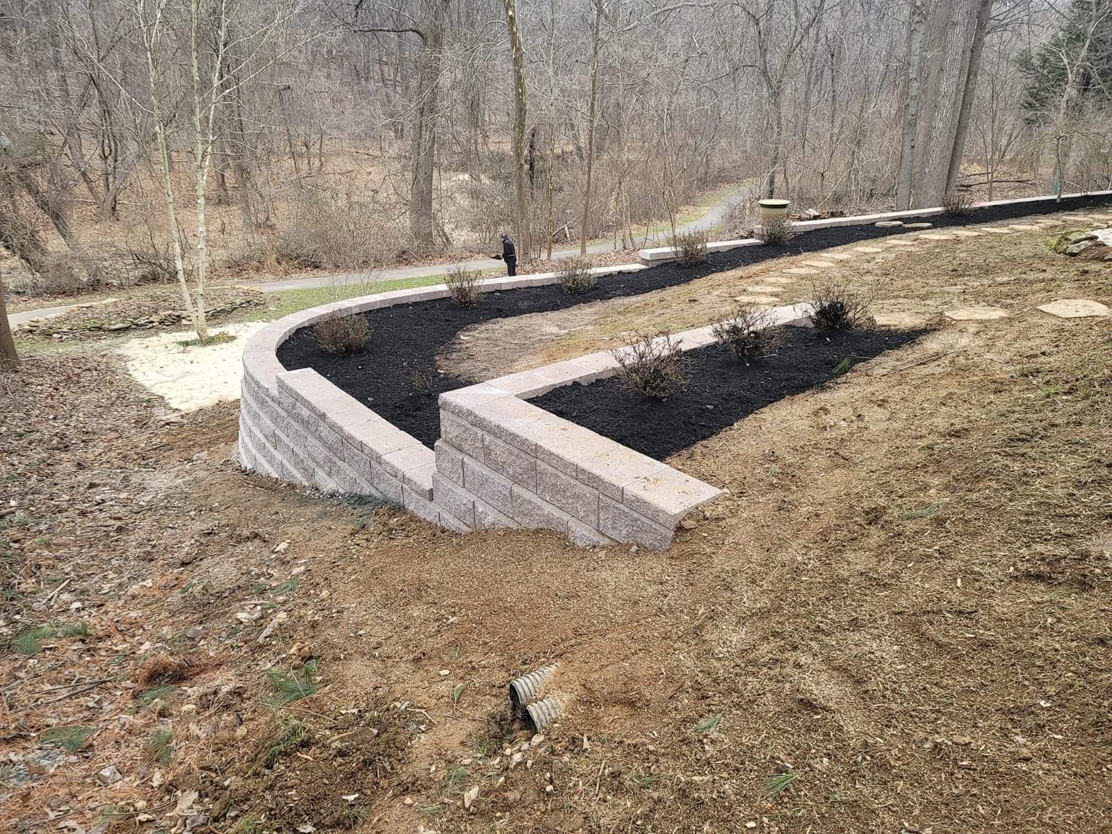 A tiered retaining wall with a curved section and steps, filled with mulch and plants, on a hillside.
