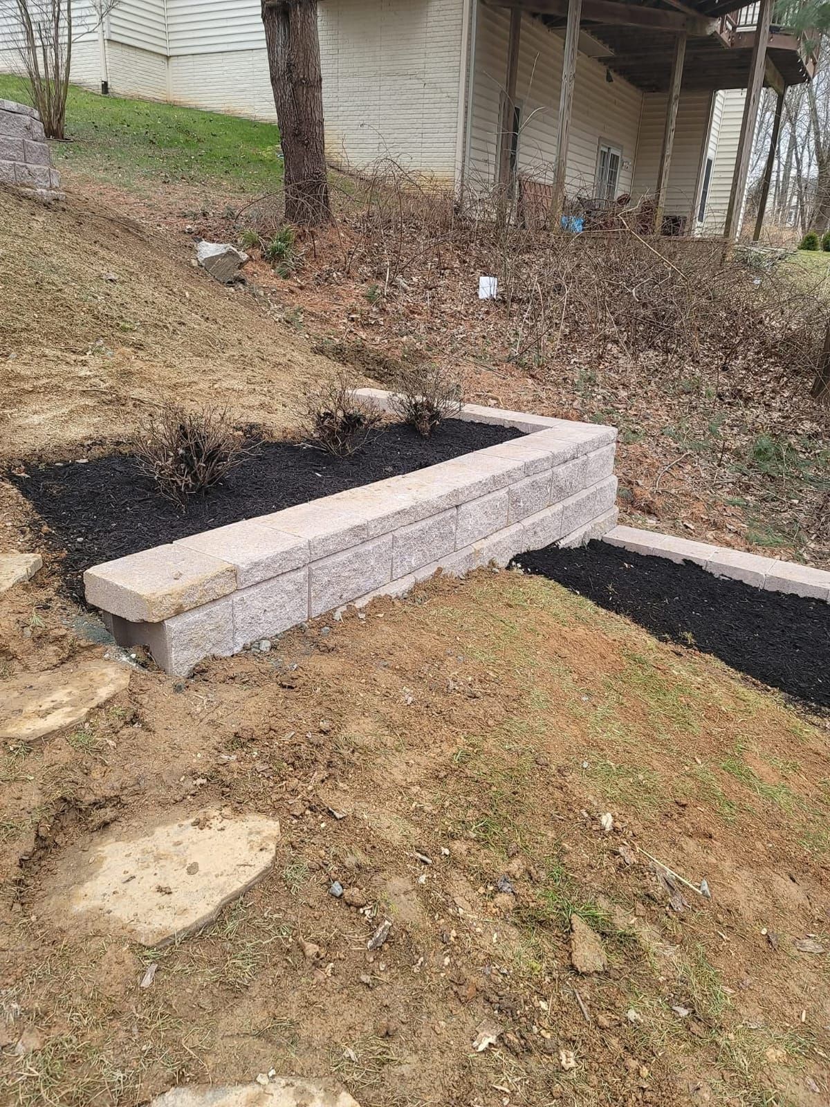 Retaining wall with dark mulch, soil, and stepping stones on a grassy hill near a house.