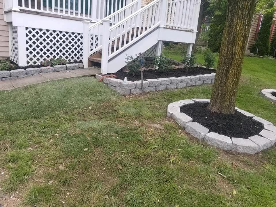 A white staircase leads to a house with a small stone-edged garden bed. A tree has a similar stone edging.