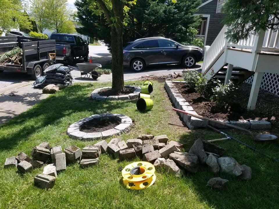 Yard work in progress: trailer, car, bricks, rocks, edging, wheelbarrow, and mulch near a house.