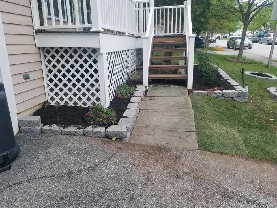 Concrete walkway leading to wooden stairs under a white deck with lattice. Landscaping with dark mulch and gray block edging.