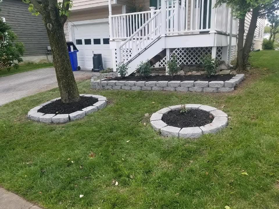 A home with a white porch and steps. Lawn with two tree rings and a flower bed, all edged with gray blocks.
