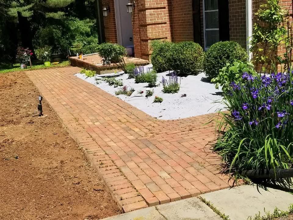 Brick walkway leading to a house with landscaping; white stones, bushes, and purple flowers.