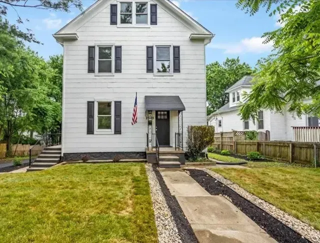 White two-story house with black shutters, awning, and American flag. Green grass and trees surround it.