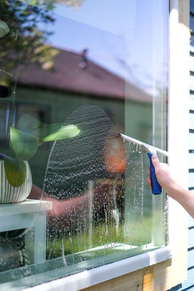 Hand using a squeegee to clean a soapy window, plants and house visible in the reflection.
