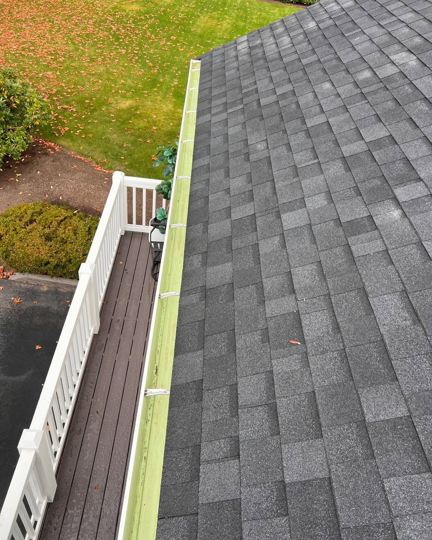 Roof of a house with gray shingles, a green gutter, and a white railing. Fall foliage in the background.