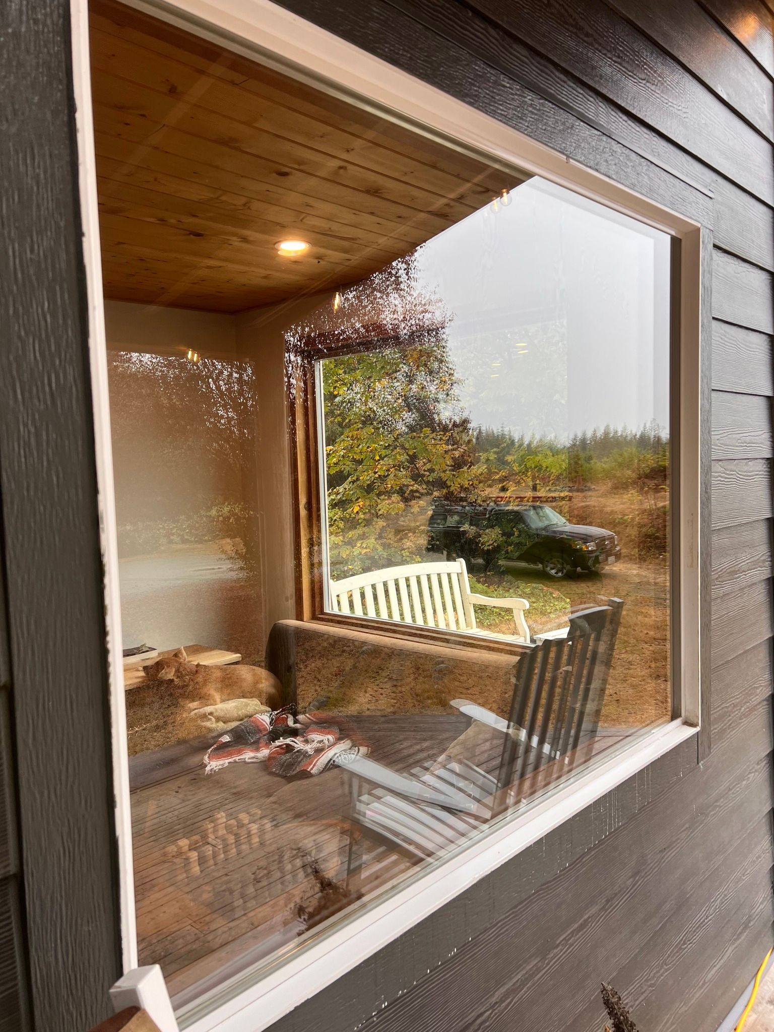 A window view. Outside, a vehicle, and chairs; inside, wooden ceiling and exposed wall, with reflection.