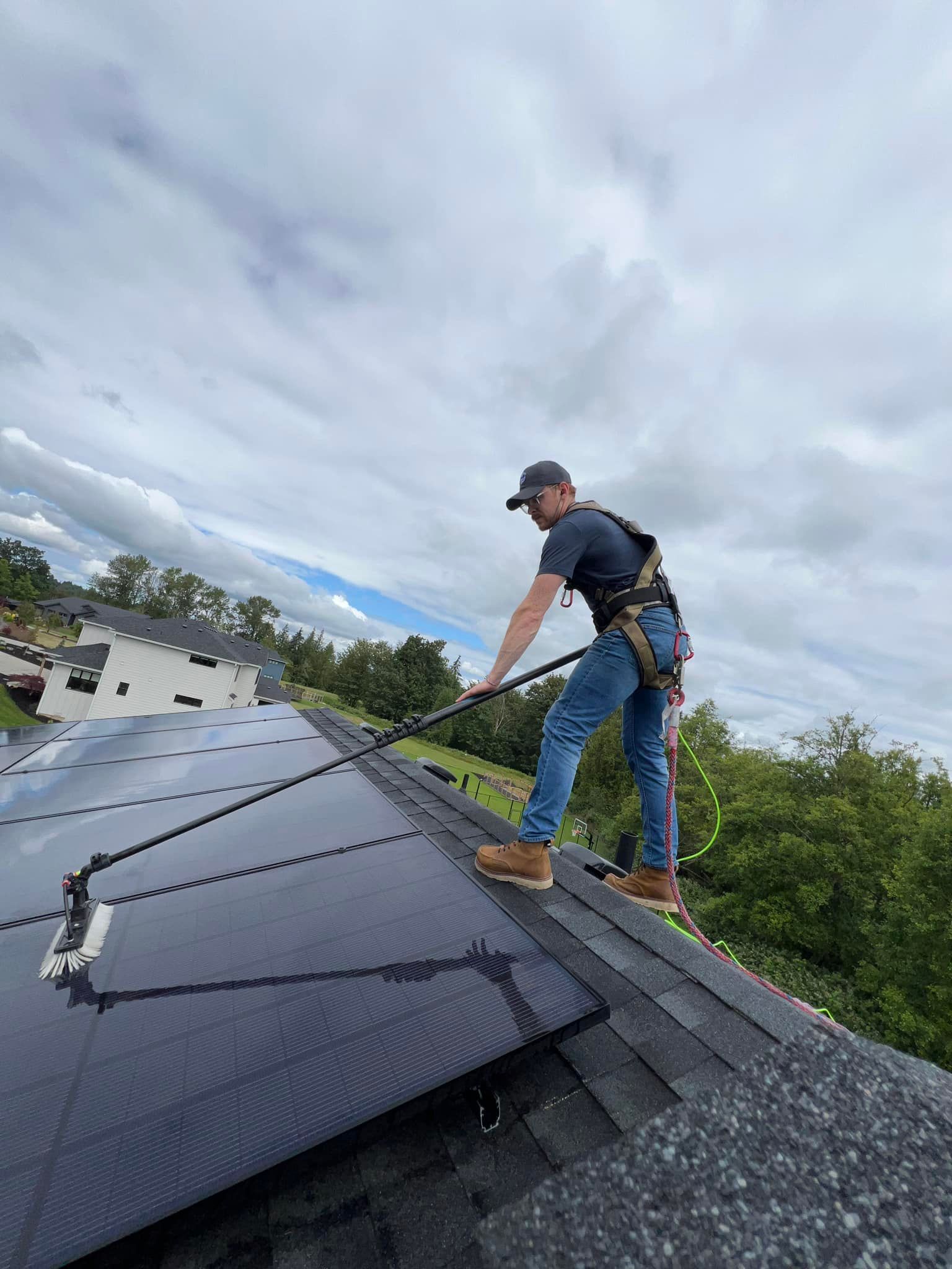 Person on roof cleaning solar panels with a long-handled brush, wearing a safety harness. Overcast sky.