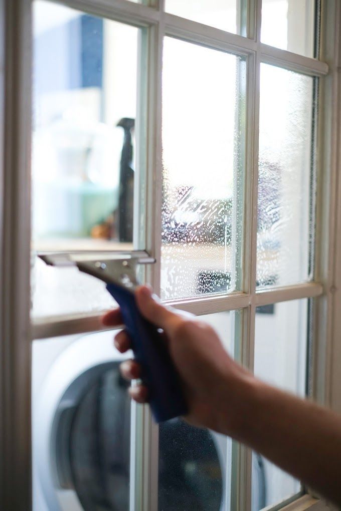 Hand using a squeegee to clean a glass window with white trim, laundry room in the background.