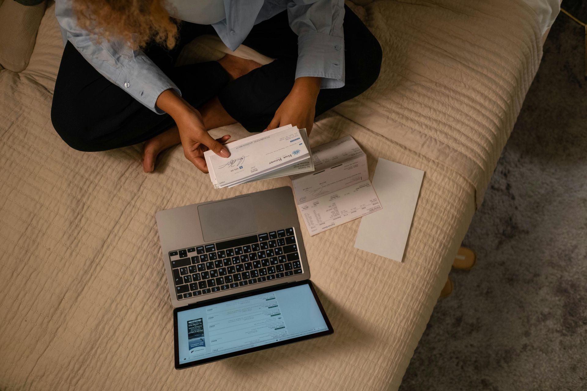 A woman is sitting on a bed with a laptop and papers.