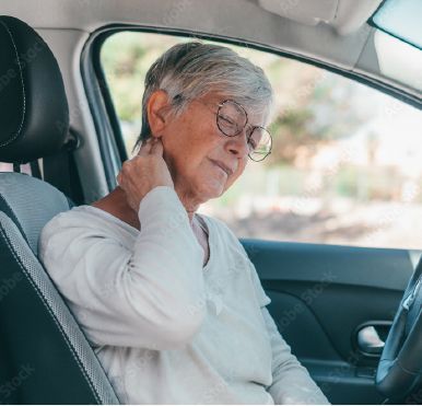 An elderly woman wearing glasses is sitting in the driver 's seat of a car holding her neck in pain.