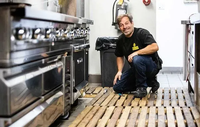 Man kneels in kitchen, smiling, next to stove and wooden floor grates.