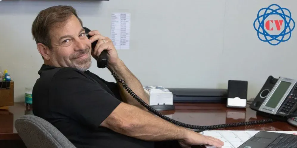 Man on the phone at his desk, smiling. Office setting with phone, desk, and logo in background.