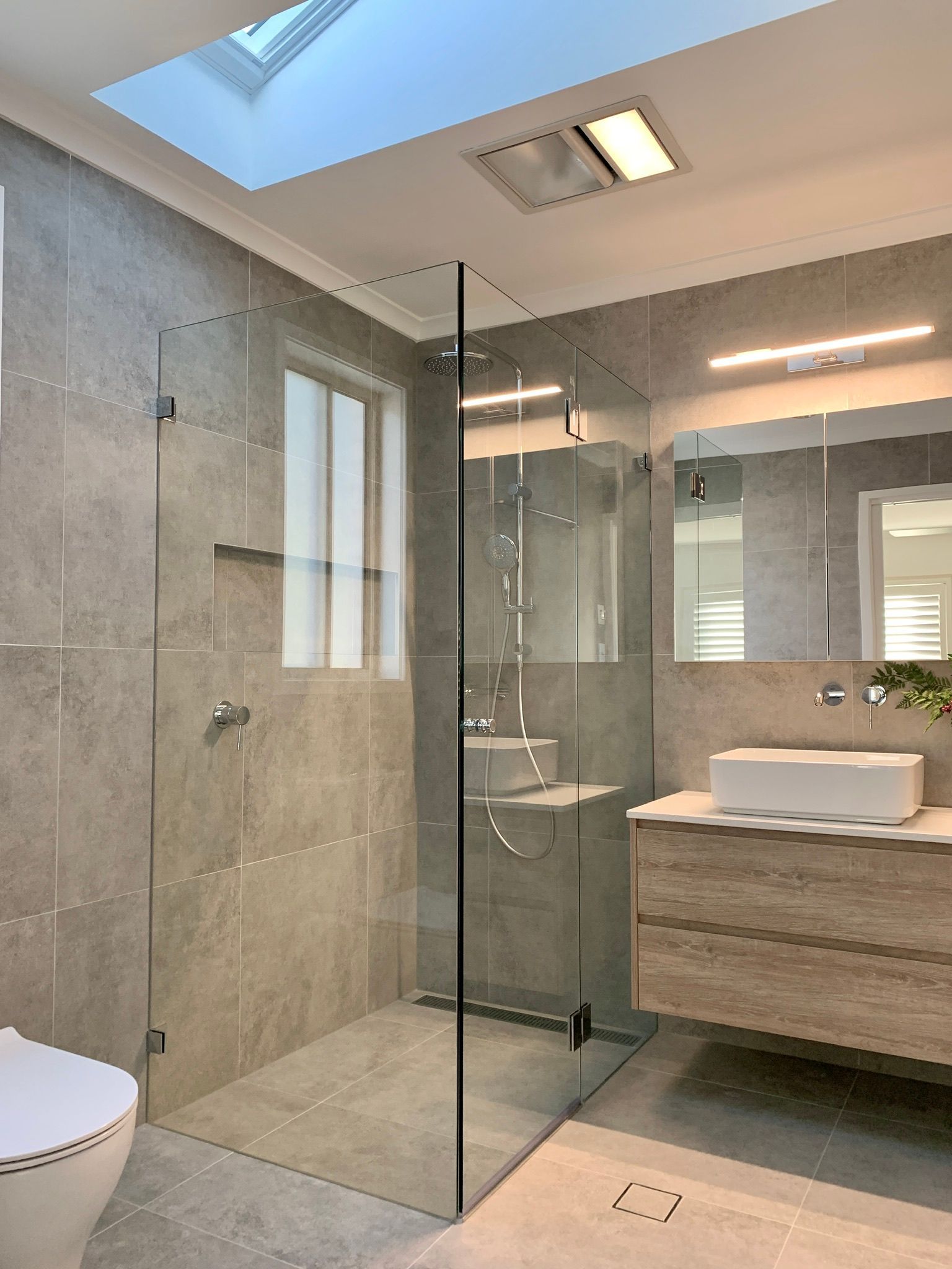 Modern bathroom with glass shower, gray tiles, skylight, and wooden vanity.