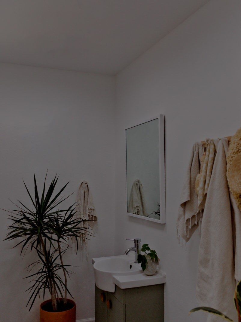 A modern bathroom with white walls, a sink, mirror, and a potted plant. Beige towels hang on the wall.
