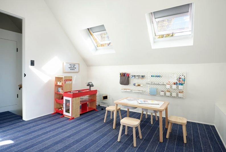 Playroom with angled ceiling, skylights, toy kitchen, small table, stools, pegboard, and blue striped rug.
