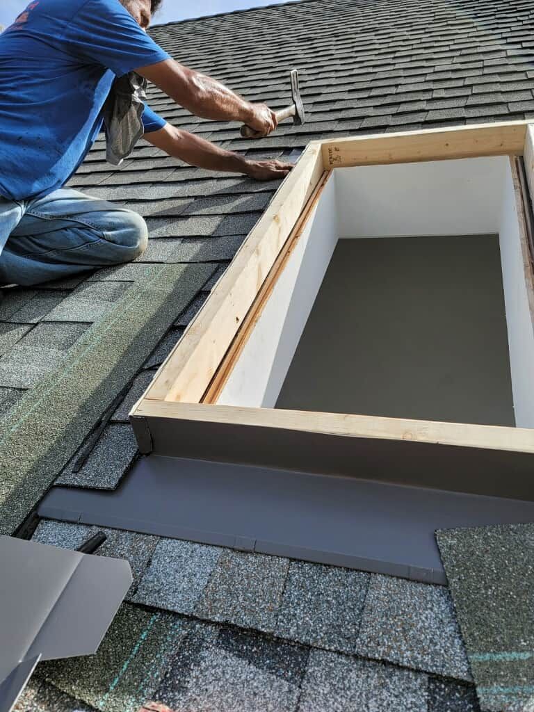 Roofer installing shingles around a skylight. A wooden frame surrounds the skylight opening.