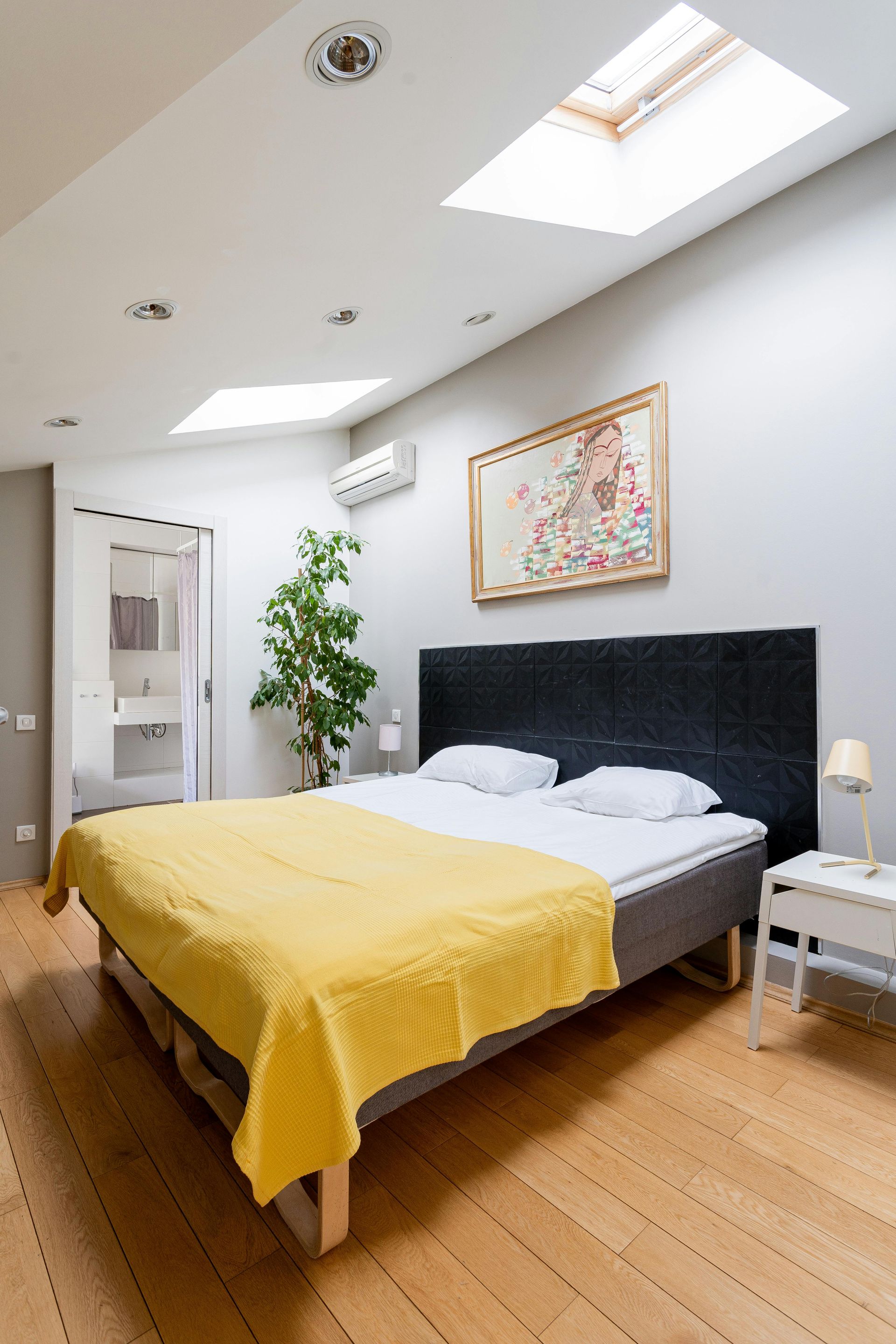 Bedroom with bed, yellow blanket, artwork, skylights, wooden floor, and a doorway to a bathroom.