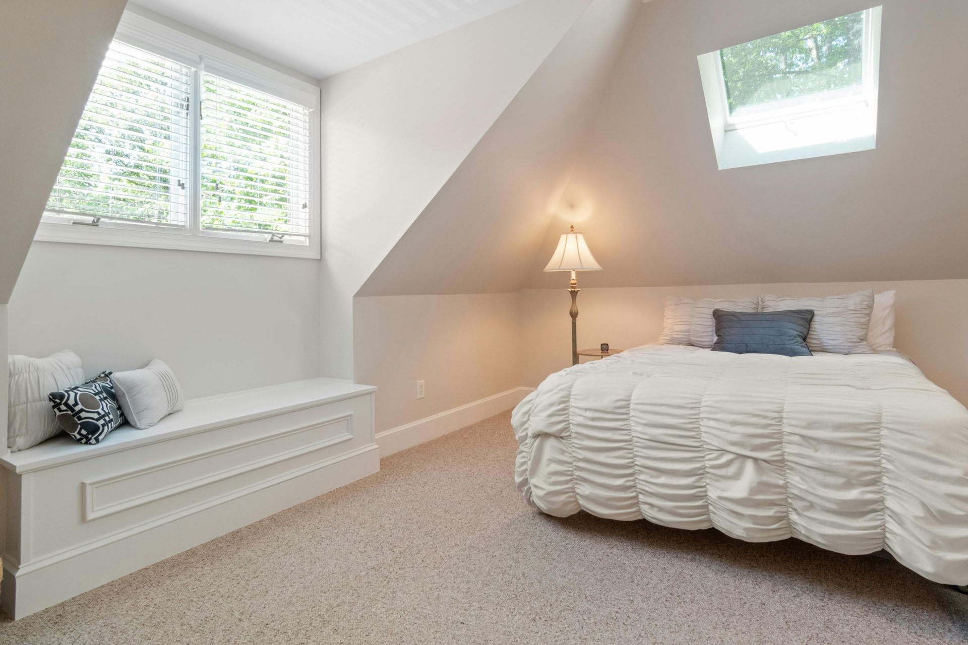 Cozy attic bedroom with bed, window seat, and skylight; neutral color scheme.