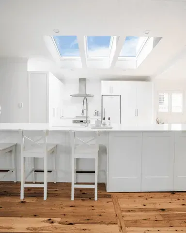 White modern kitchen with skylights, island, wooden floor, and bar stools.