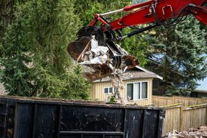 A red excavator is loading a dumpster with a house in the background.