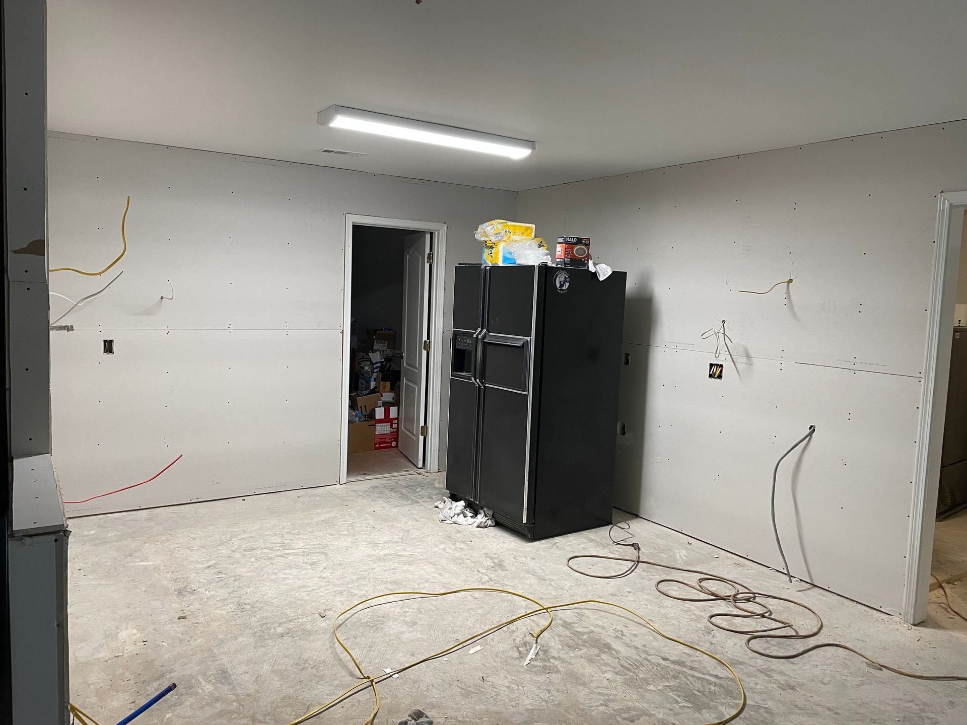 Basement room with unfinished drywall, exposed wiring, and a black refrigerator near an open doorway.
