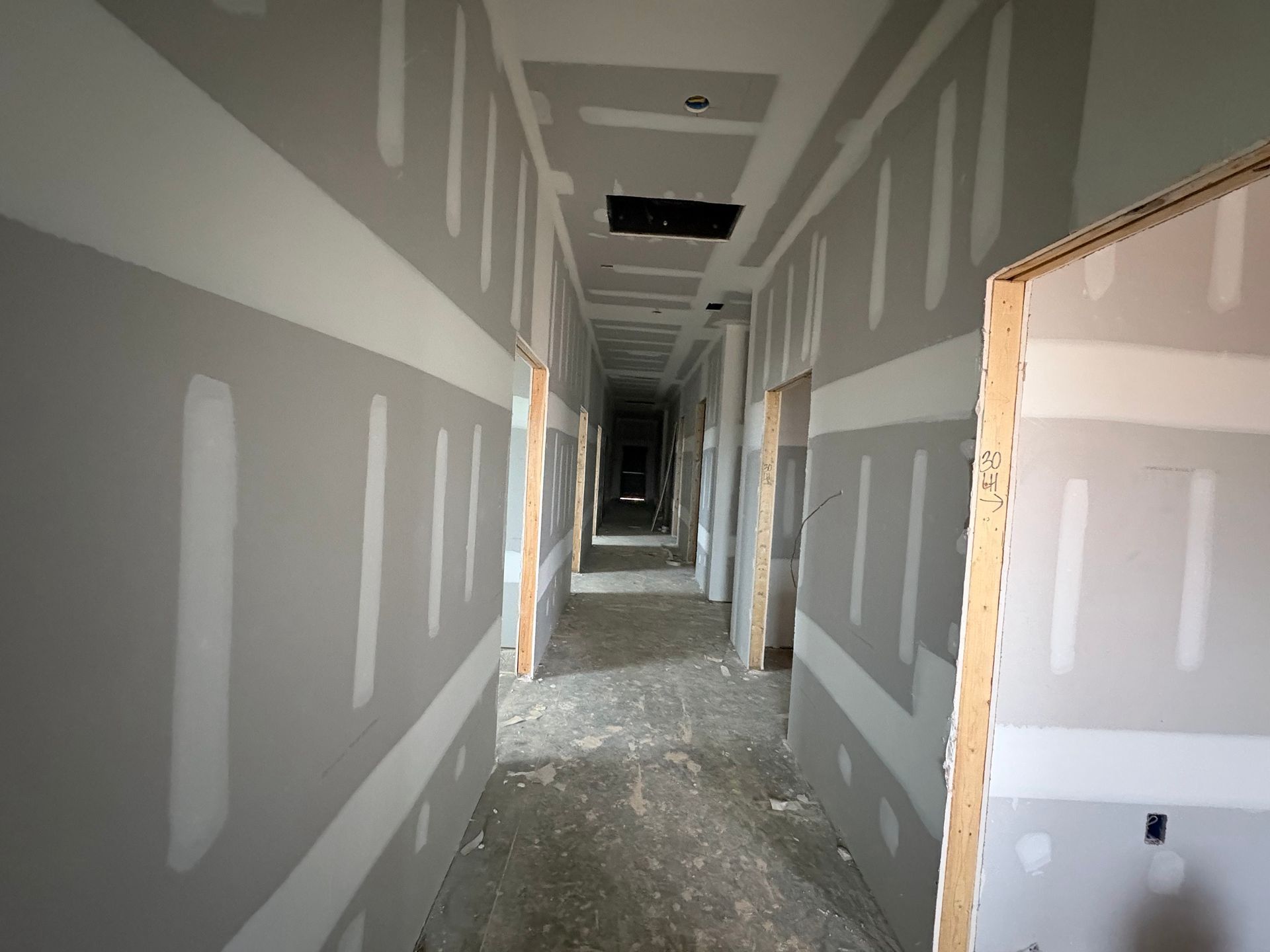 Long unfinished hallway with gray drywall, exposed ceiling, and construction debris on the floor