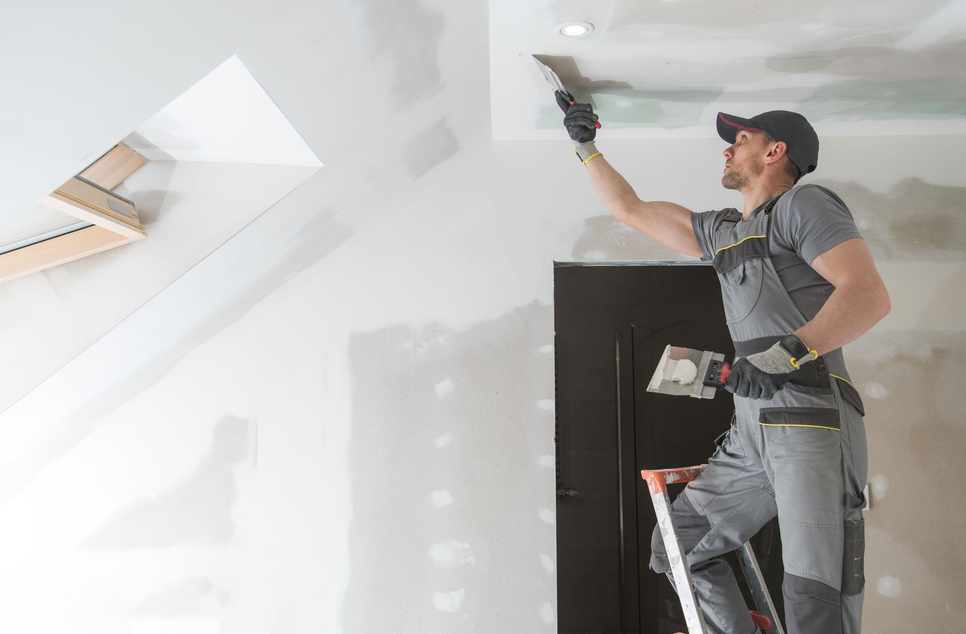 Worker using a drill to install a ceiling fixture in a white room, standing on a ladder.