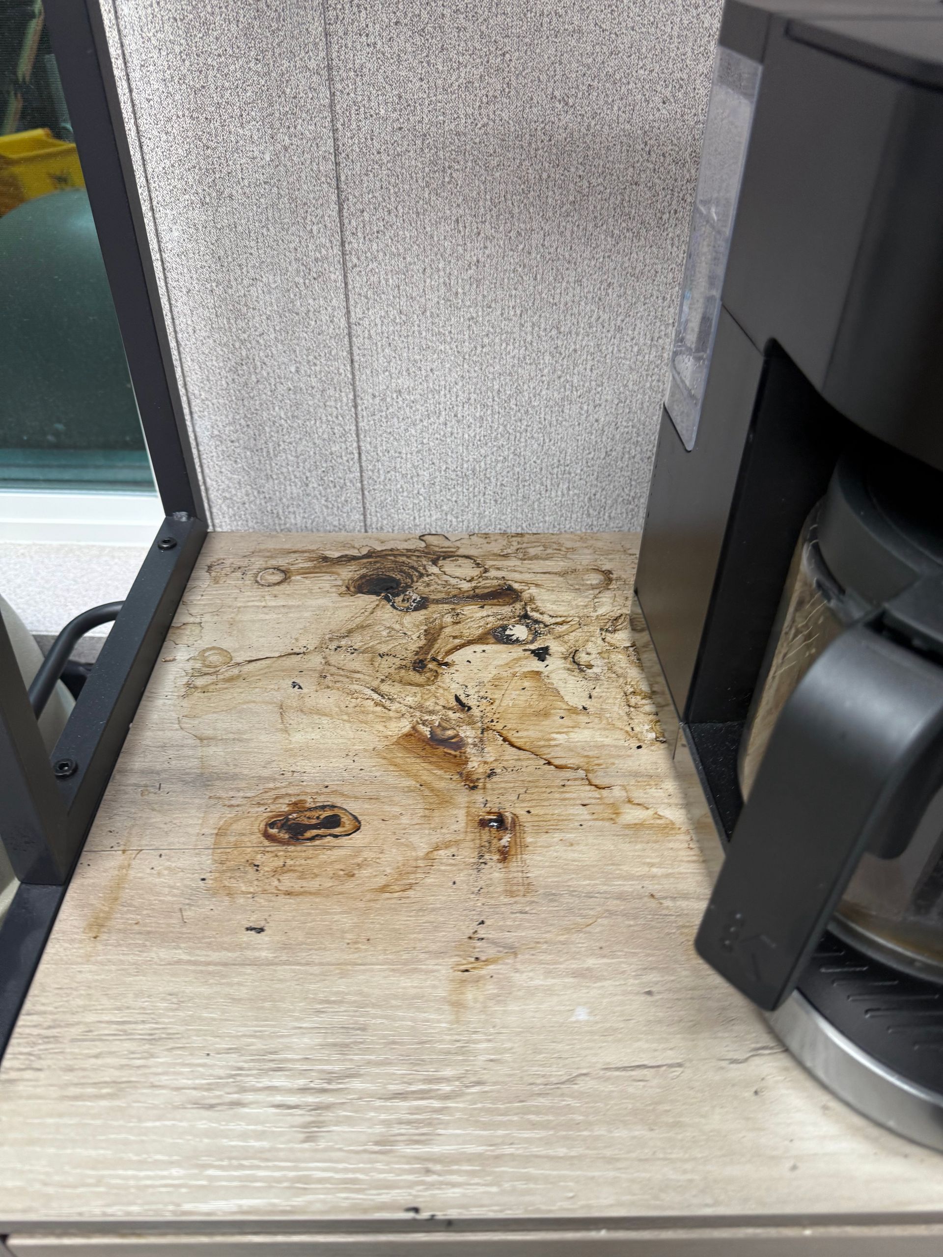 Coffee maker on a wooden shelf stained with coffee spills against a neutral wall.