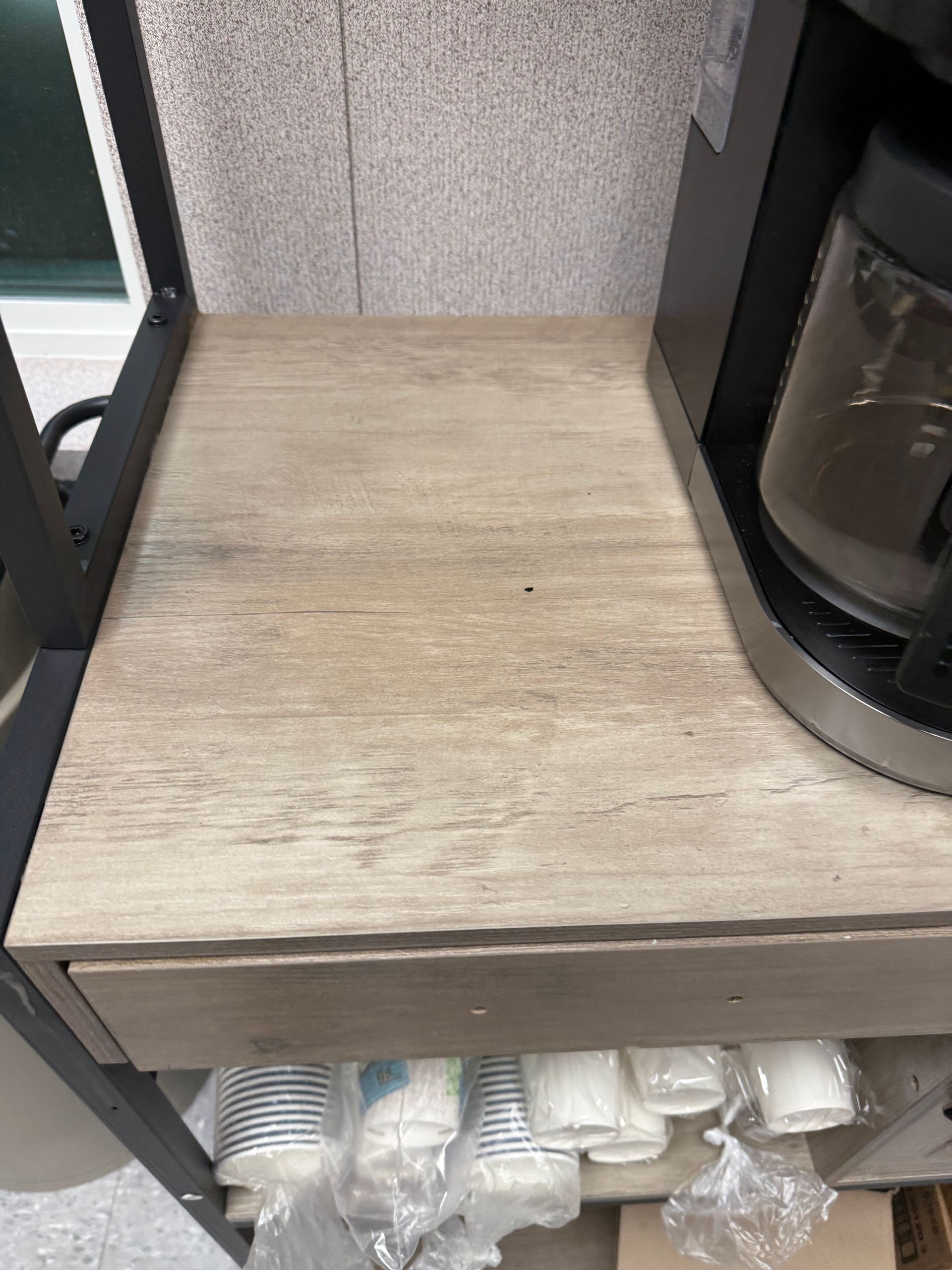 Coffee maker on a shelf. Cups below. The shelf has a wood-like finish. Gray background.