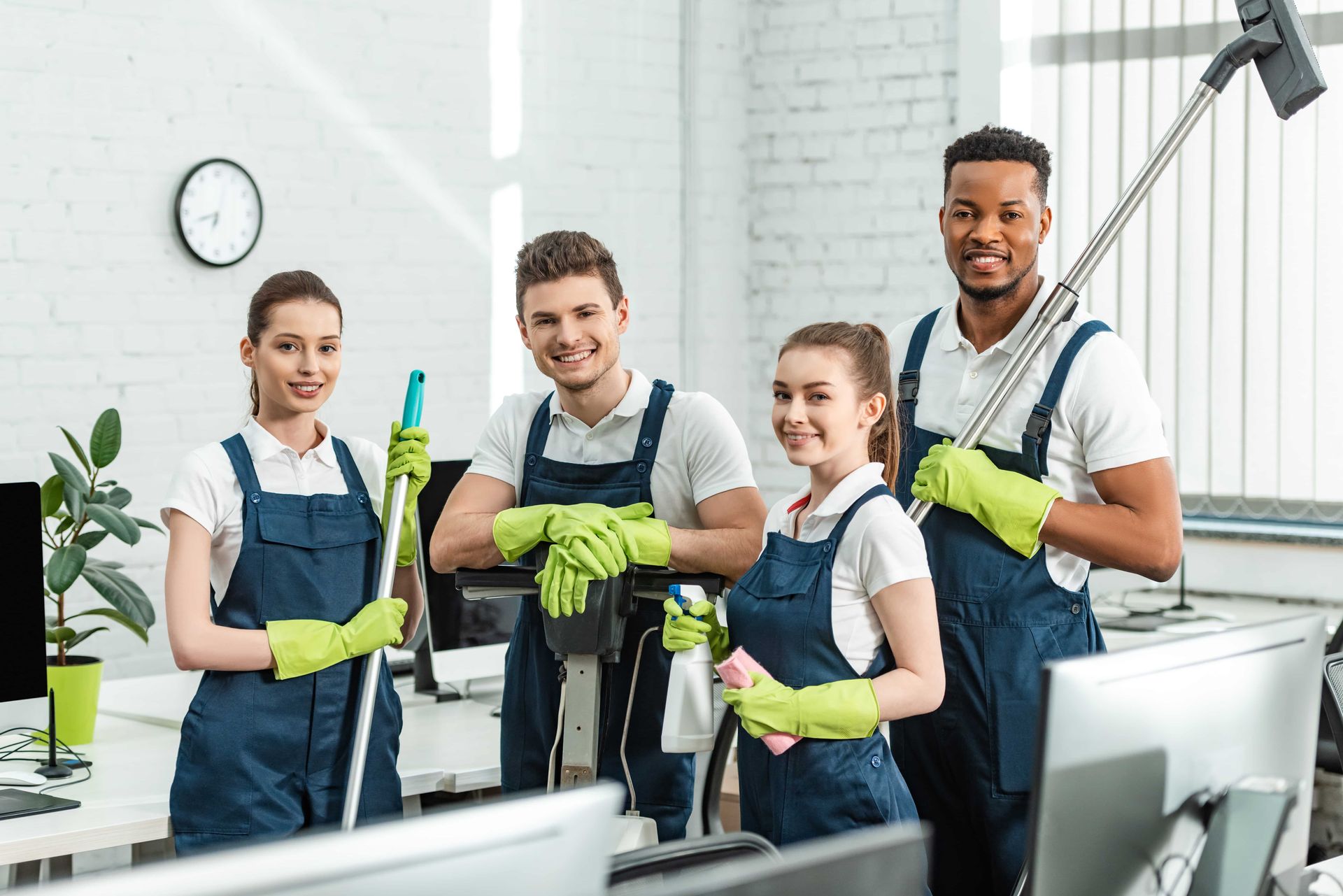 Four smiling office cleaners in blue aprons and green gloves, holding cleaning supplies in a bright office.