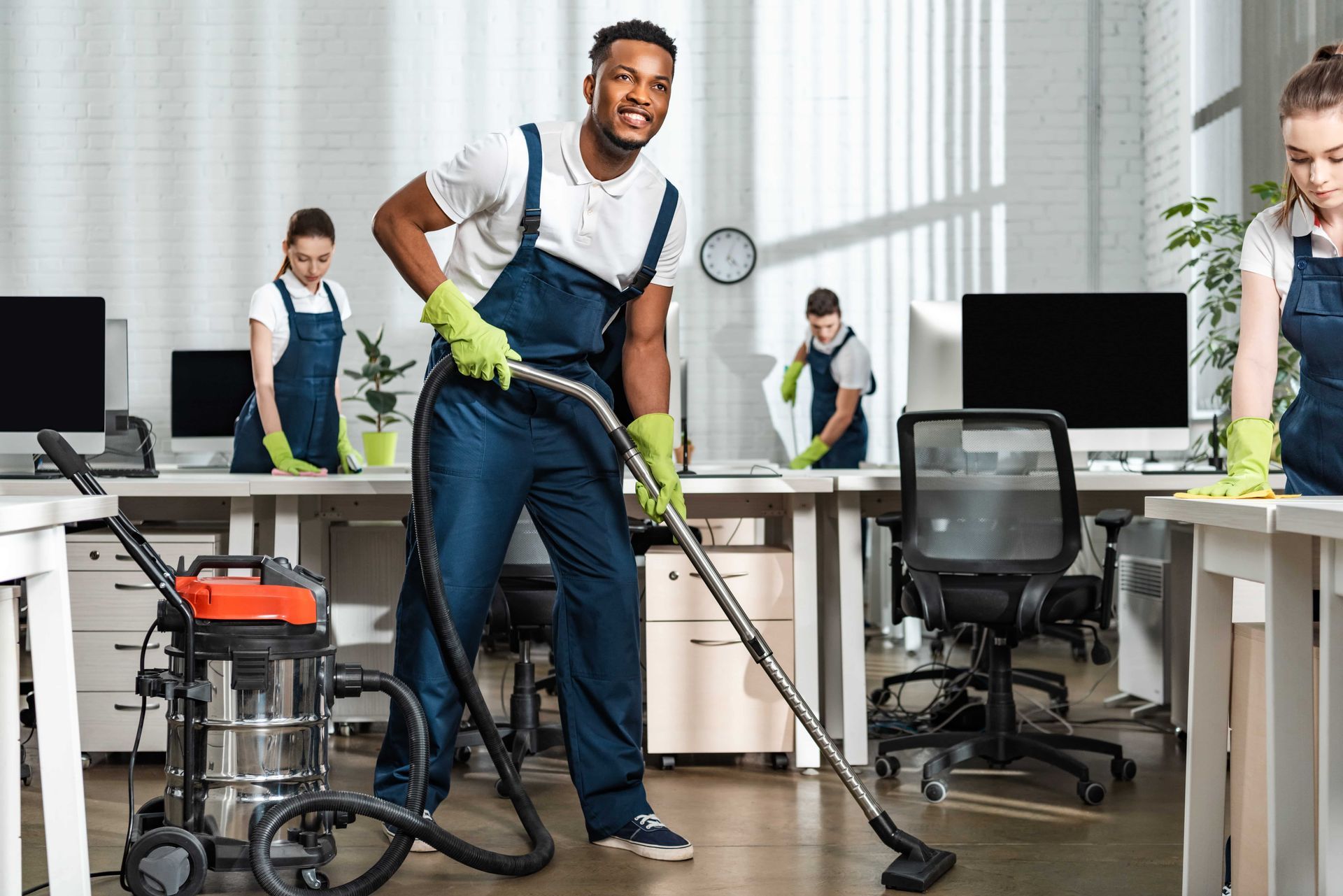 A smiling Black man in a blue uniform vacuums an office; two others clean desks.